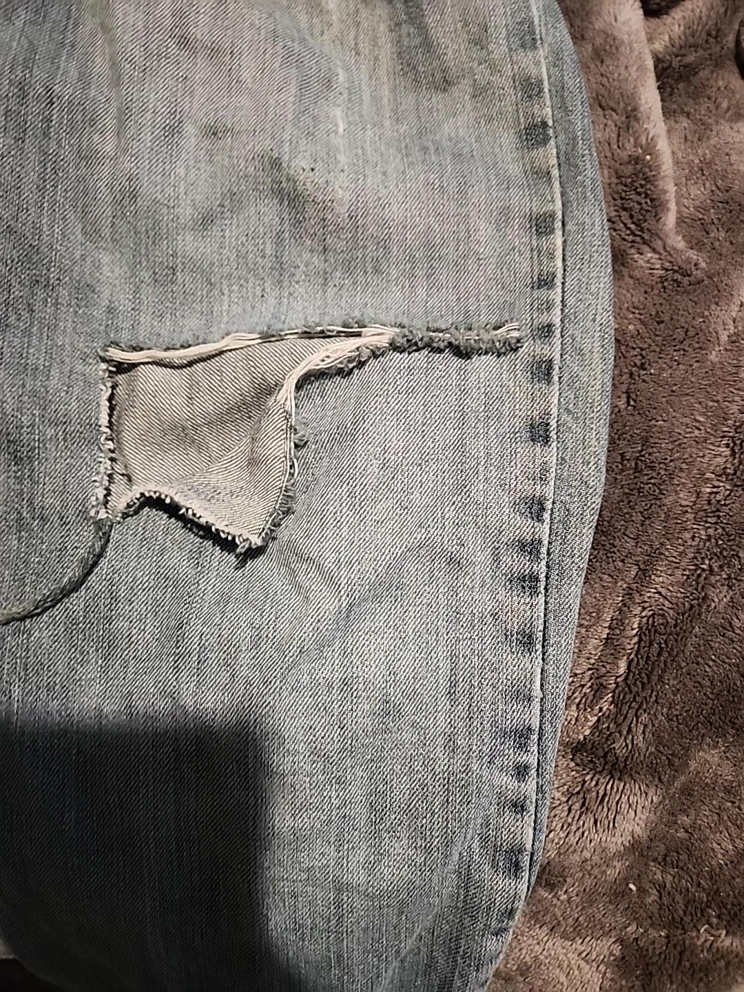 Faded jeans with a frayed split beside the outseam. White weft threads bridge a horizontal tear, and a flap of denim has dropped open to show the pale reverse.